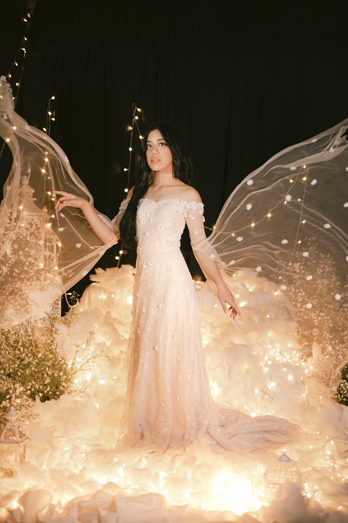 A stunning woman in an elegant dress stands amidst a fairy-tale setting with glowing lights and ethereal fabric.
