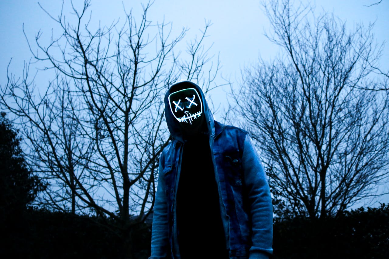Eerie image of a person with LED mask and hoodie standing among bare trees at twilight.